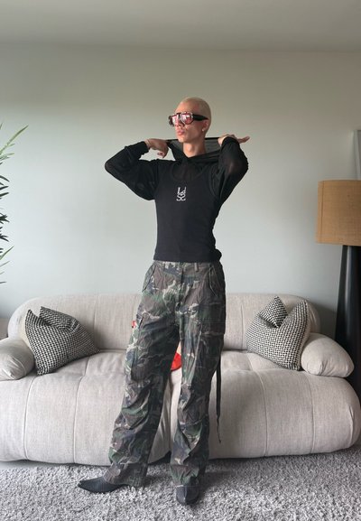 Camisa de malla negra de manga larga superpuesta a una camiseta negra ajustada, combinada con pantalones cargo de camuflaje. El modelo lleva gafas de sol rojas de gran tamaño.