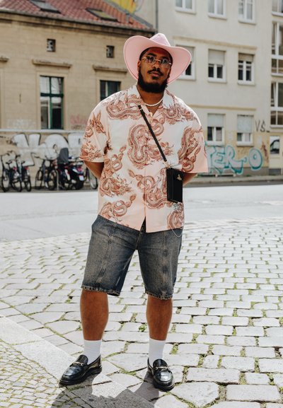 Camisa de estilo kimono en rosa con patrón de dragón rojo; pantalones cortos de mezclilla; mocasines negros; calcetines blancos; sombrero de ala ancha rosa; gafas de sol.