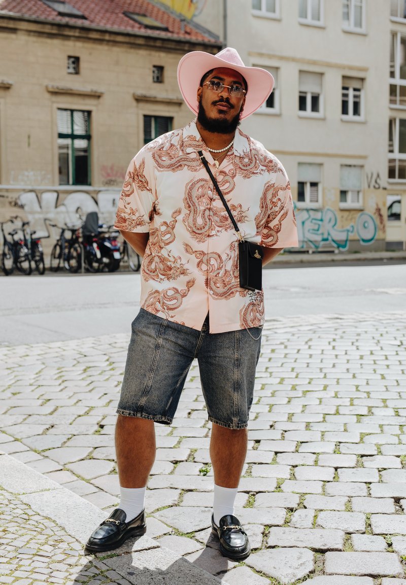 Πουκάμισο στυλ κιμονό σε ροζ με σχέδιο κόκκινου δράκου; τζιν σορτς; μαύρες εσπαντρίγιες; λευκές κάλτσες; ροζ καπέλο με φαρδύ γείσο; γυαλιά ηλίου.