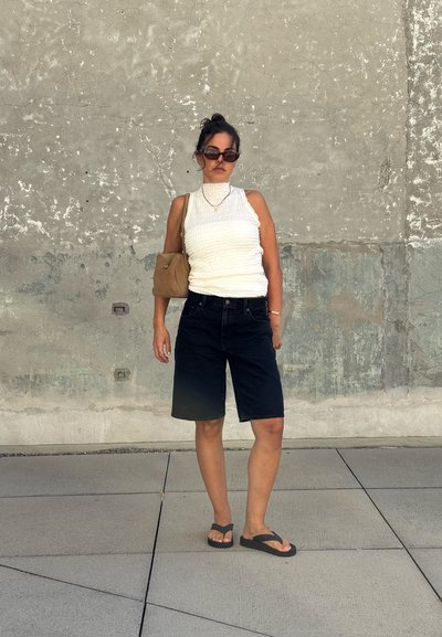 Top sin mangas de textura blanca, pantalones cortos de mezclilla oscuros a la altura de la rodilla, chanclas negras, bolso marrón, de pie contra una pared gris texturizada.