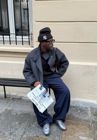 Grijze wollen jas over een zwarte trui, navy wijde broek, en zilveren sneakers; houdt een krant vast terwijl ze op een zwart bankje zit.