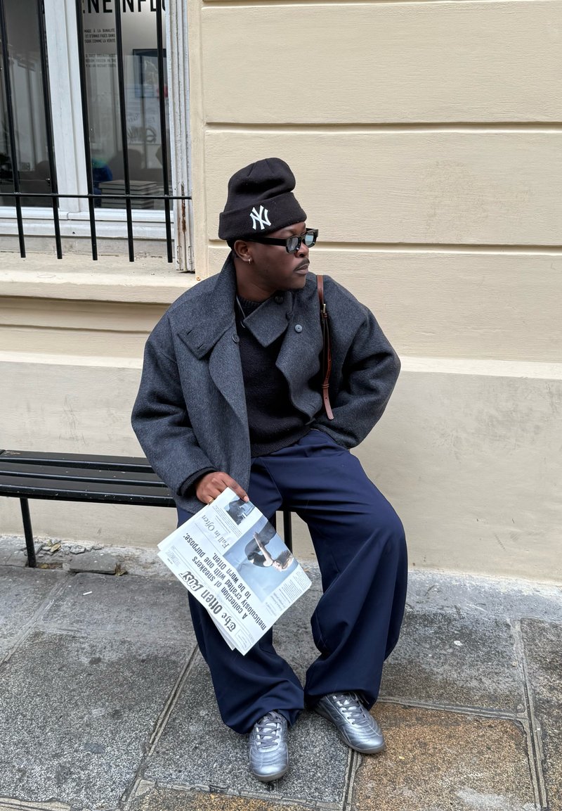 Manteau en laine gris par-dessus un pull noir, pantalon large bleu marine, et baskets argentées ; tenant un journal tout en étant assis sur un banc noir.