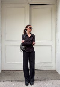 Camisa preta de botão, calças pretas largas, sapatos de plataforma pretos e óculos de sol escuros. Bolsa crossbody com tachas, encostada a uma porta branca.