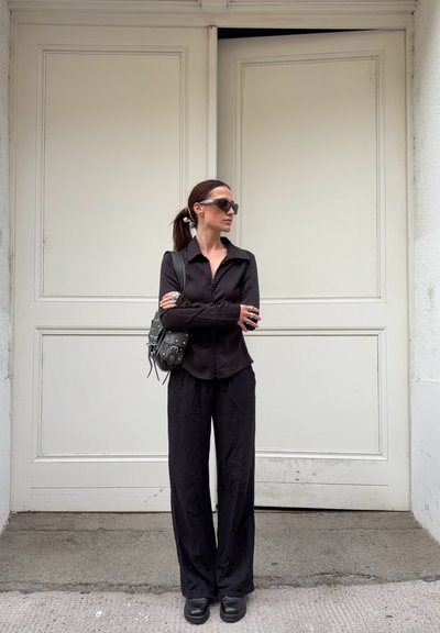 Camisa negra de botones, pantalones negros holgados, zapatos de plataforma negros y gafas de sol oscuras. Bolsa cruzada con tachuelas, de pie contra una puerta blanca.