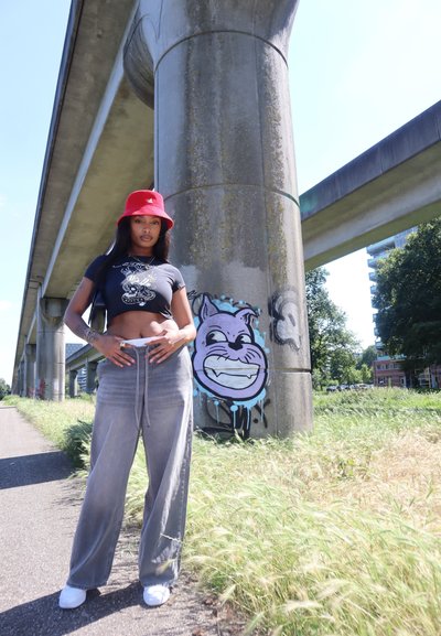 Sombrero de cubo rojo, camiseta gráfica negra ajustada, pantalones de pierna ancha gris claro, zapatos blancos. El fondo presenta un pilar de concreto y graffiti.