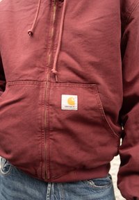 Μαρουλί zip-up φούτερ φτιαγμένο από βαμβάκι, με μπροστινή τσέπη και λευκή ετικέτα με πορτοκαλί λογότυπο. Το ύφασμα φαίνεται υφασμένο και ανθεκτικό.