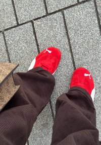 Pieds portant des baskets Puma rouges et blanches et un pantalon en velours côtelé marron, debout sur un trottoir en pierre grise.