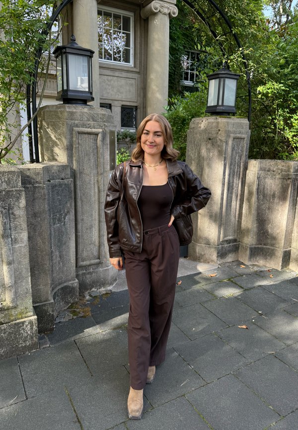 Braune Lederjacke, schwarzes Oberteil, lockere braune Hose und beige Knöchelstiefel. Außenszene mit Steinsäulen und Grünpflanzen im Hintergrund.