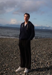 Une personne se tient sur une plage de galets, portant un sweat à capuche bleu marine, un pantalon noir et des baskets blanches, avec des montagnes et de l'eau en arrière-plan.