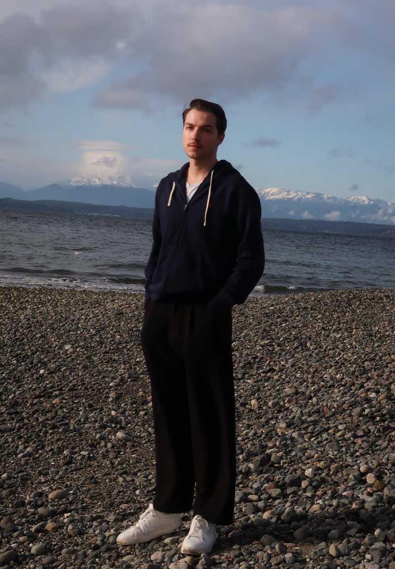 Een persoon staat op een kiezelstrand, gekleed in een marineblauwe hoodie, zwarte broek en witte sneakers, met bergen en water op de achtergrond.