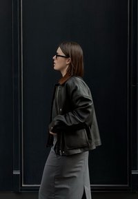 Melnā ādas jaka ar brūnu apkakli, uzvilkta virs pelēka apģērba. Dizainam ir brīva piegriezne un izteiksmīgas šuvju detaļas.