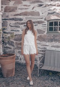 Hvit ermeløs topp med utskjæringer og shorts med bølgekant, parret med slip-on sko, mot en steinvegg.