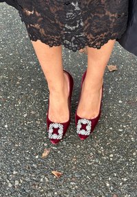 Décolleté in raso bordeaux con un ornamento quadrato di strass sulla punta, caratterizzate da una forma arrotondata e una texture liscia. Sfondo con pavimento visibile.