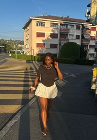 Chemise noire à manches courtes, jupe évasée blanche et sandales noires ; portant un petit sac. Marchant sur un passage piéton avec un bâtiment en arrière-plan.
