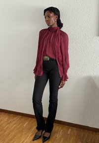Mørkerød, struktureret bluse med voluminøse ærmer, parret med tætsiddende sorte bukser og spidse sorte hæle. Sølvspændt bælte fremhæver taljen.