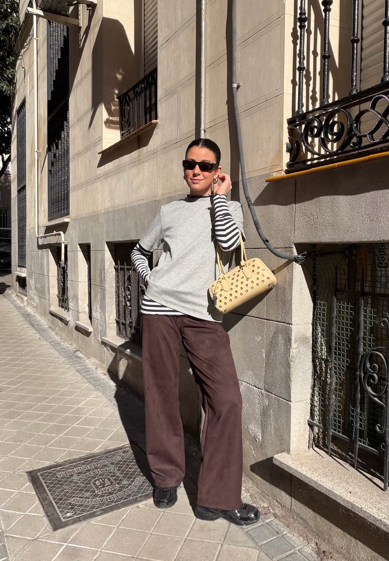Mujer con gafas de sol negras, camiseta de manga corta gris sobre mangas largas a rayas, pantalones marrones y sosteniendo un bolso beige con tachuelas en una calle soleada.