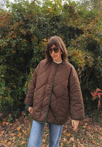 Barna steppelt dzseki zöld részletekkel, gallér nélküli kialakítással és két elülső zsebbel. Kék farmernadrág felett viselve, zöld növényzet előtt.