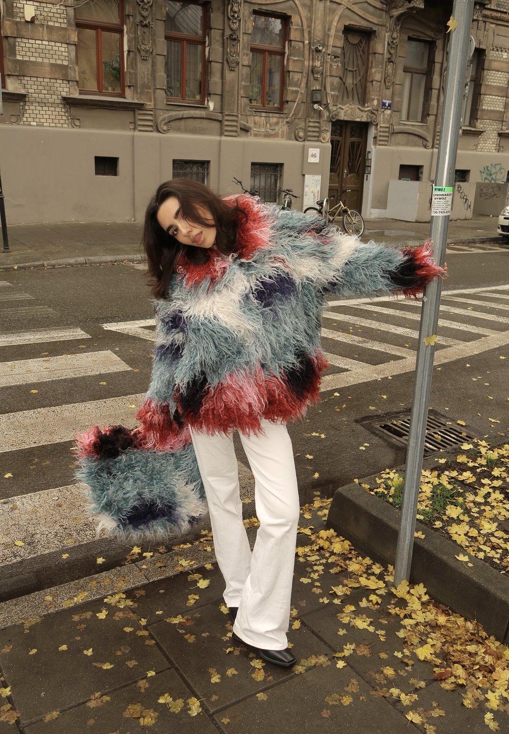 Többszínű, piros, kék, fehér és fekete bolyhos pulóver hozzávaló kézitáskával. A modell fehér bővülő nadrágot visel, az utcán áll.