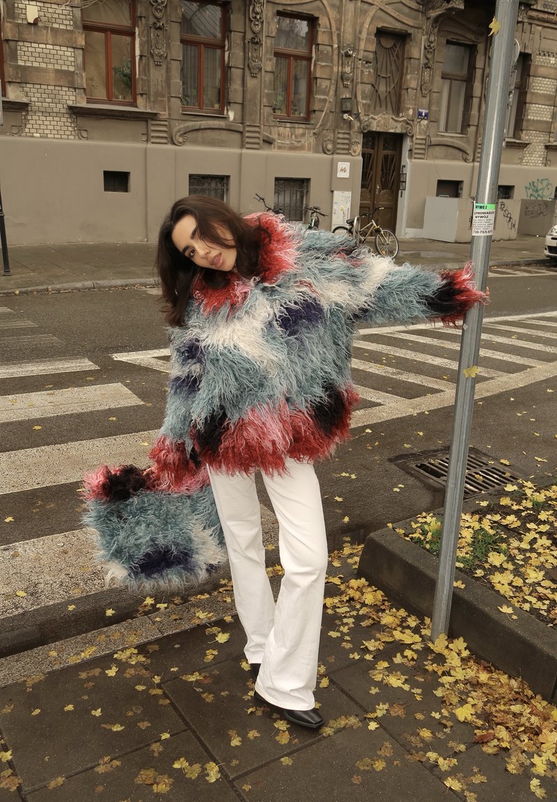 Multicoloured shaggy jumper in red, blue, white and black with a matching handbag. Model wears white flared trousers, standing on a street.