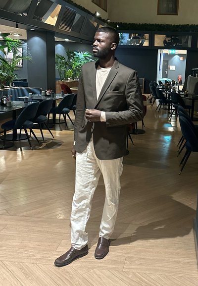 Blazer marrón a medida sobre una camisa beige, combinado con pantalones de color claro y zapatos marrones. Ambiente interior con plantas y decoración moderna.