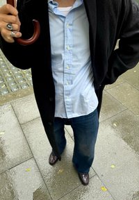 Isik kannab musta pintsakut, helesinist särke, siniseid teksaseid ja pruune kingi, hoides pruunist vihmavarjust niiskel sillutisel, kuhu on langenud lehti.