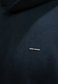 Σκούρος ναυτικός φούτερ φτιαγμένο από μαλακό ύφασμα, με μία μικρή ορθογώνια μαύρη ετικέτα στην οποία είναι ραμμένο το όνομα "UNDER ARMOUR."