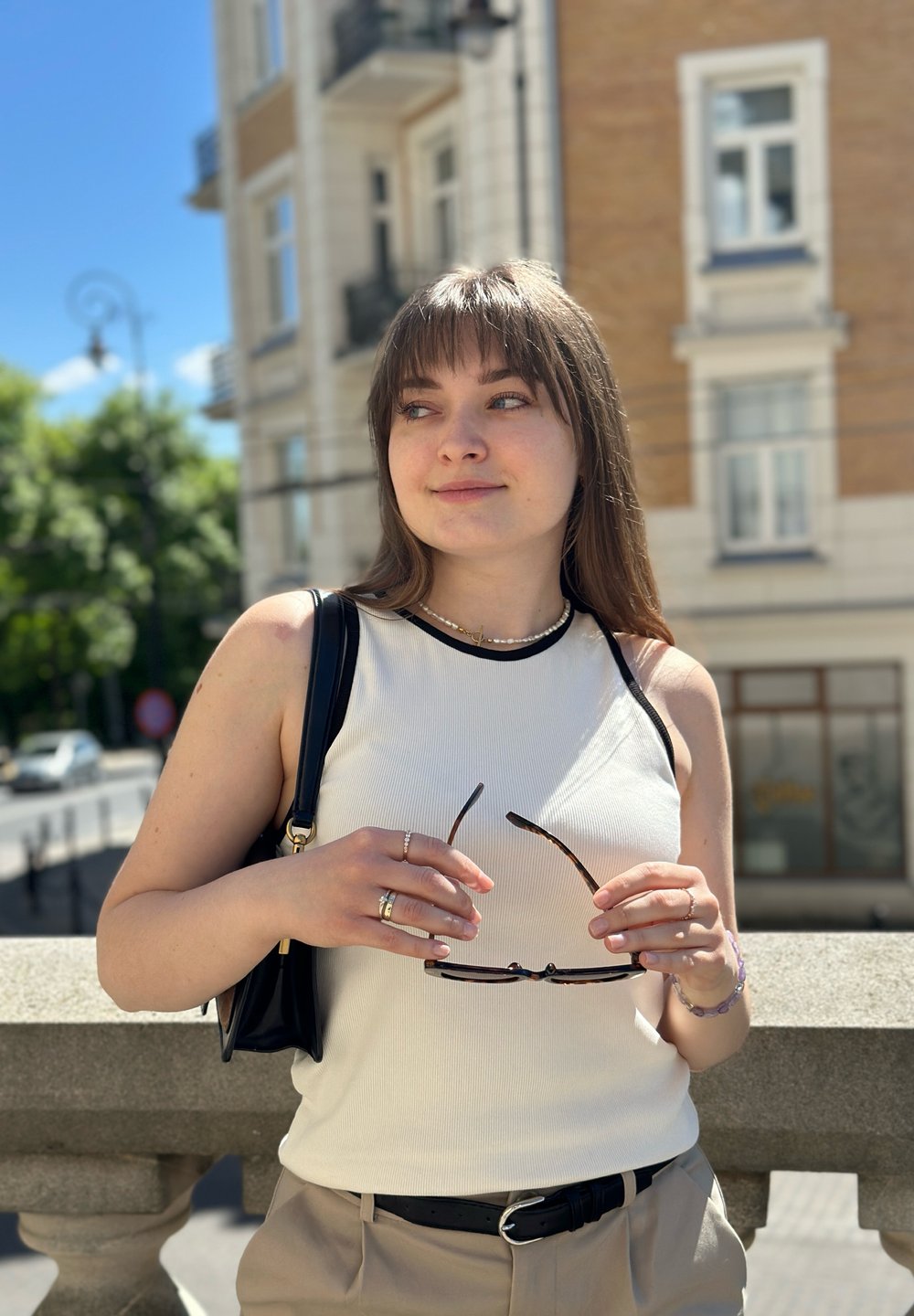 Nainen, joka pitää käsissään ruskeita aurinkolaseja, on pukeutunut valkoiseen hihattomaan toppiin, jossa on mustia yksityiskohtia, vaaleisiin housuihin ja mustaan olkalaukkuun. Taustalla auringonvalossa kylpevä kaupunkimaisema.