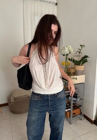 Jeune femme aux longs cheveux bruns, portant un haut blanc à nouer derrière le cou et un jean large bleu, ajustant un sac à bandoulière noir à l'intérieur près de plantes.