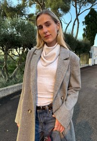 Manteau long à motif pied-de-poule gris, haut à col boule crème, jean taille haute, ceinture métallique, tenant un sac bordeaux, en extérieur avec des arbres en arrière-plan.