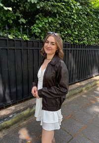 Jovem mulher a sorrir, vestindo um vestido branco, uma jaqueta de couro castanho, com óculos de sol na cabeça, de pé ao lado de uma cerca de metal preta com folhagem verde atrás.