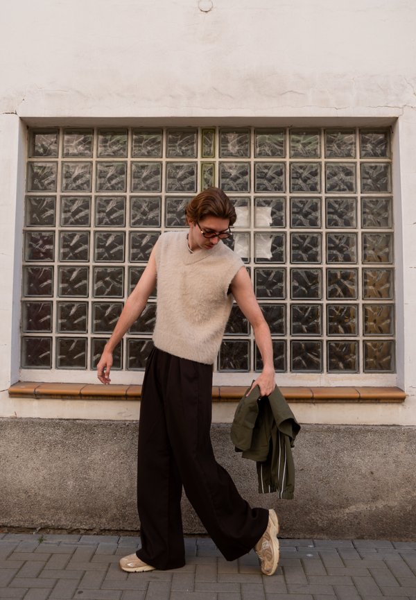 Persona joven que lleva un suéter beige sin mangas y pantalones anchos negros, sostiene una chaqueta verde, de pie en una acera frente a bloques de vidrio.