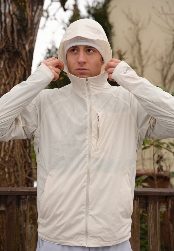 Junger Mann im Freien, der die Kapuze seiner hellbeigen, wasserdichten Jacke mit hohem Kragen und Reißverschlusstasche auf der Brust justiert.