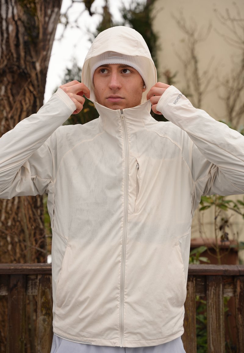 Junger Mann im Freien, der die Kapuze einer hellbeigen, wasserdichten Jacke mit hohem Kragen und Reißverschlusstasche auf der Brust anpasst.