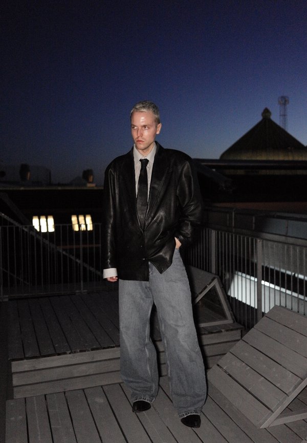 Homme portant une veste en cuir noire, un jean gris à jambes larges et des chaussures noires, debout sur une terrasse sur le toit au crépuscule, les mains dans les poches.