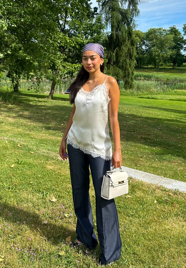 Woman with long dark hair wears a white lace camisole, dark jeans, headscarf, and holds a white handbag, standing in a sunny park.