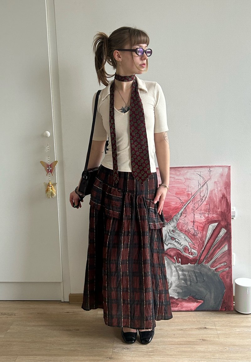 Femeie cu coadă de cal, purtând ochelari, bluză crem, eșarfă cu modele, fustă lungă în carouri și pantofi negri, stând în interior lângă o pictură cu un dragon.