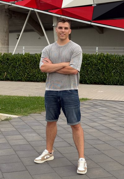 Camisa de punto de manga corta gris claro con rayas verticales, pantalones cortos de mezclilla azul y zapatillas deportivas de color claro con acentos beige, de pie sobre el pavimento.