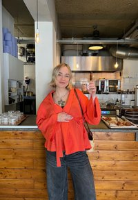 Κόκκινο, υφασμένο, μακρυμάνικο τοπ τύπου περιτυλίγματος με ζώνη στη μέση, συνδυασμένο με φαρδιά, σκούρα τζιν. Το φόντο περιλαμβάνει μια ξύλινη επιφάνεια και εξοπλισμό κουζίνας.