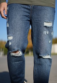 Jeans in denim blu stressato con strappi visibili alle ginocchia e bordi sfrangiati, caratterizzati da una vestibilità rilassata e una leggera scoloritura.