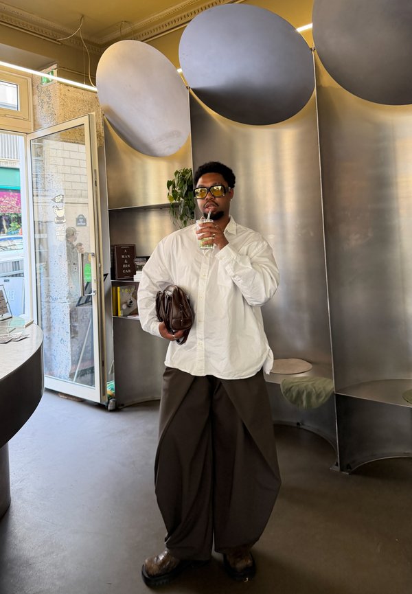 Man in wit hemd en brede donkere broek houdt een bruine tas vast en drinkt met een rietje in een modern café met metalen inrichting.