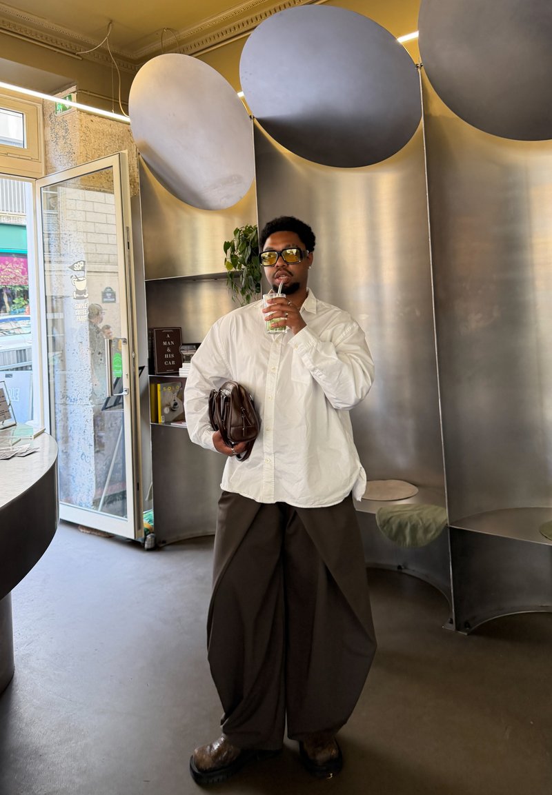 Homme en chemise blanche et pantalon large foncé tenant un sac marron et buvant à la paille dans un café moderne avec une décoration métallique.