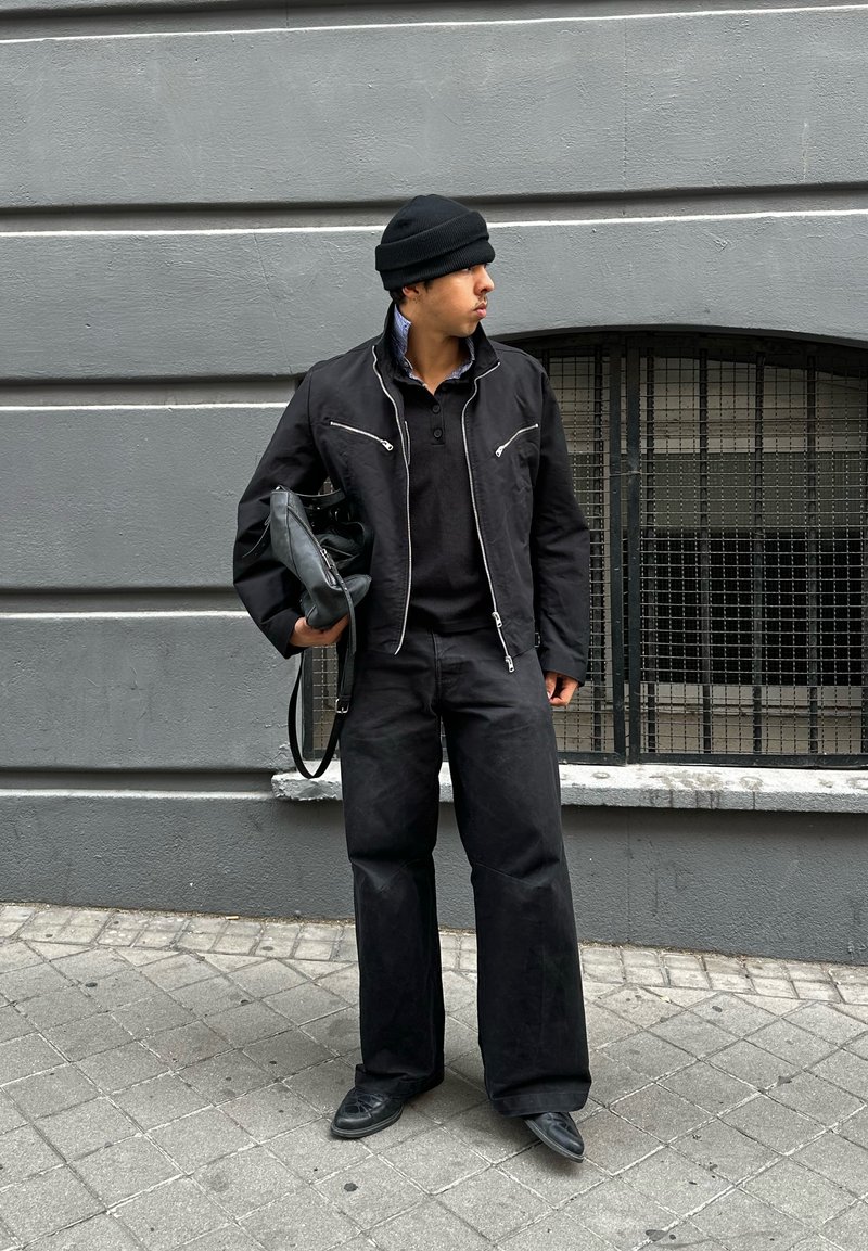 Casaco preto com fecho éclair e costuras brancas contrastantes, um suéter preto por baixo e calças pretas largas. A segurar uma bolsa preta, de pé sobre o pavimento.