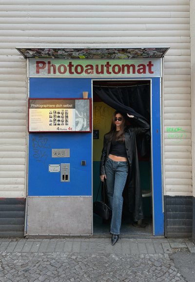 Mujer con gafas de sol y abrigo largo negro saliendo de una cabina de fotos azul y blanca con la etiqueta "Photoautomat" en una acera urbana.