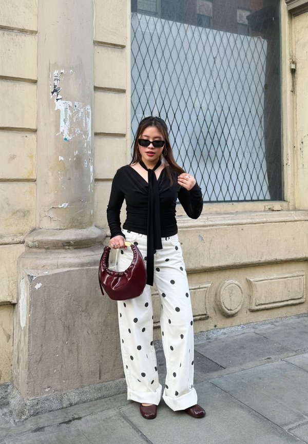 Frau steht auf dem Bürgersteig in der Stadt, trägt ein schwarzes Oberteil mit Schaldetail, weiße Polka-Dot-Hose, Sonnenbrille, burgunderfarbene Schuhe und hält eine burgunderfarbene Handtasche.