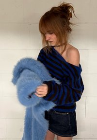 Ženska z rdečimi lasmi v nepravilno speti figi, oblečena v modro-črni črtasti pulover z odkritimi rameni in jeans krilo, drži veliko puhasto svetlo modro ruto.