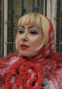 Czerwona dzianinowa szal z białym, kratkowanym wzorem, ułożona na puszystym różowo-szarym płaszczu. Jaskrawy niebieski makijaż oczu i wyraziste czerwone usta.