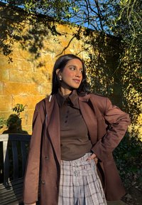 Manteau brun croisé sur un polo marron foncé. Jupe à carreaux gris avec bretelles bouclées. Décor naturel avec un mur en pierre en arrière-plan.