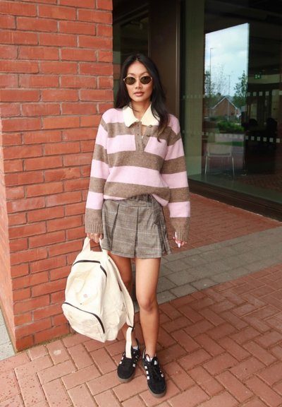 Mujer joven con gafas de sol, suéter de rayas, falda de cuadros, zapatillas negras, sosteniendo una mochila blanca, de pie junto a una pared de ladrillos fuera de un edificio.