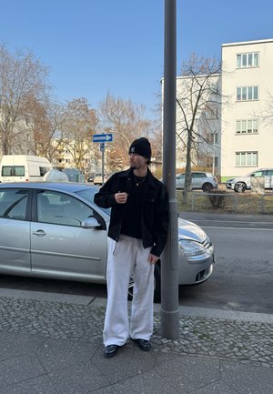 Chaqueta de mezclilla negra sobre una camiseta negra, pantalones de chándal blancos y zapatos negros. Lleva un gorro negro y está de pie junto a un coche plateado.