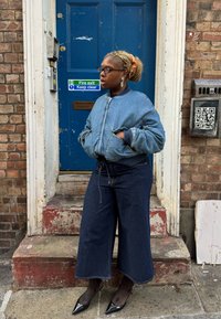 Denim bomberjakke med taljeindsnævring, vidde bukser i mørk denim og spidse sorte hæle. Håret er sat op i en hestehale med en orange scrunchie.